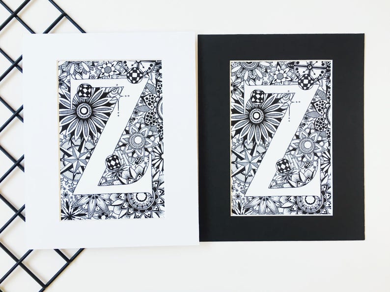May include: Two framed prints of a black and white illustration of the letter Z surrounded by intricate patterns. The print on the left is framed in white, and the print on the right is framed in black.
