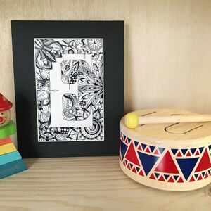 May include: A black and white framed print of the letter "E" surrounded by intricate doodle designs. The print is sitting on a wooden surface next to a wooden toy drum with a red, blue, and white pattern and a yellow drumstick.