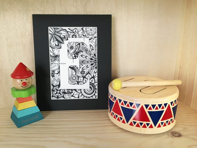 May include: A black and white framed print of the letter "E" surrounded by intricate doodle designs. The print is sitting on a wooden surface next to a wooden toy drum with a red, blue, and white pattern and a yellow drumstick.
