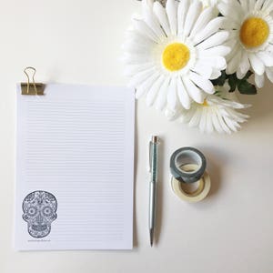 Puede incluir: Un bloc de notas rayado con un dise&ntilde;o de calavera de az&uacute;car decorativa, sujeto con un clip dorado. Un bol&iacute;grafo plateado con un cuerpo de cristal, dos rollos de cinta adhesiva decorativa y margaritas blancas tambi&eacute;n est&aacute;n presentes.