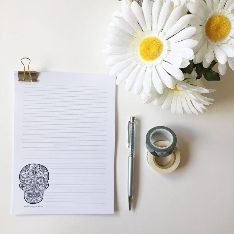 Puede incluir: Un bloc de notas rayado con un dise&ntilde;o de calavera de az&uacute;car decorativa, sujeto con un clip dorado. Un bol&iacute;grafo plateado con un cuerpo de cristal, dos rollos de cinta adhesiva decorativa y margaritas blancas tambi&eacute;n est&aacute;n presentes.