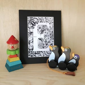 May include: A black framed print of the letter E with a black and white intricate design. The print is sitting on a wooden surface with a wooden toy clown and a set of three wooden toy penguins.