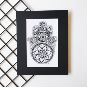 May include: A black and white framed print of a hamsa hand with an eye in the center and intricate mandala designs. The hamsa is a symbol of protection and good luck.