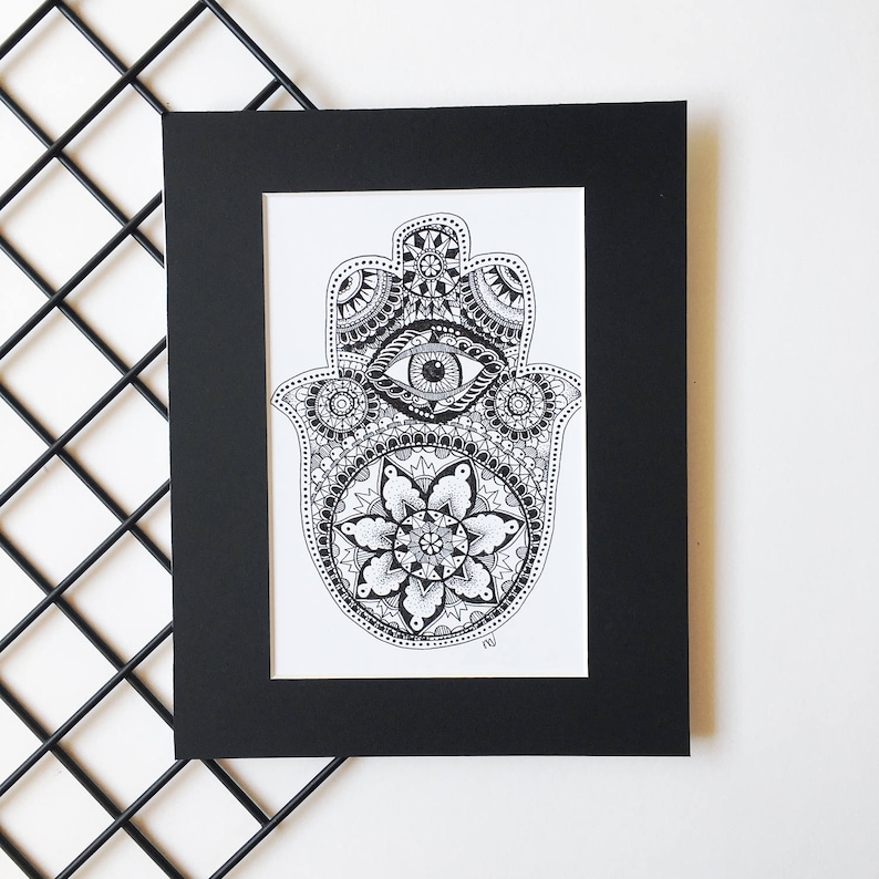 May include: A black and white framed print of a hamsa hand with an eye in the center and intricate mandala designs. The hamsa is a symbol of protection and good luck.