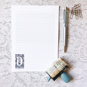 May include: A white sheet of lined paper with a black and white floral design with the letter "D" in the center. A silver pen with a sparkly blue ink cartridge and a blue and silver stamp with a variety of colorful stamps are on a white lace background.