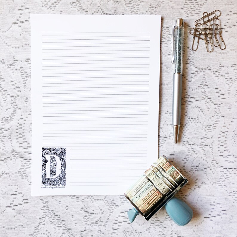 May include: A white sheet of lined paper with a black and white floral design with the letter "D" in the center. A silver pen with a sparkly blue ink cartridge and a blue and silver stamp with a variety of colorful stamps are on a white lace background.