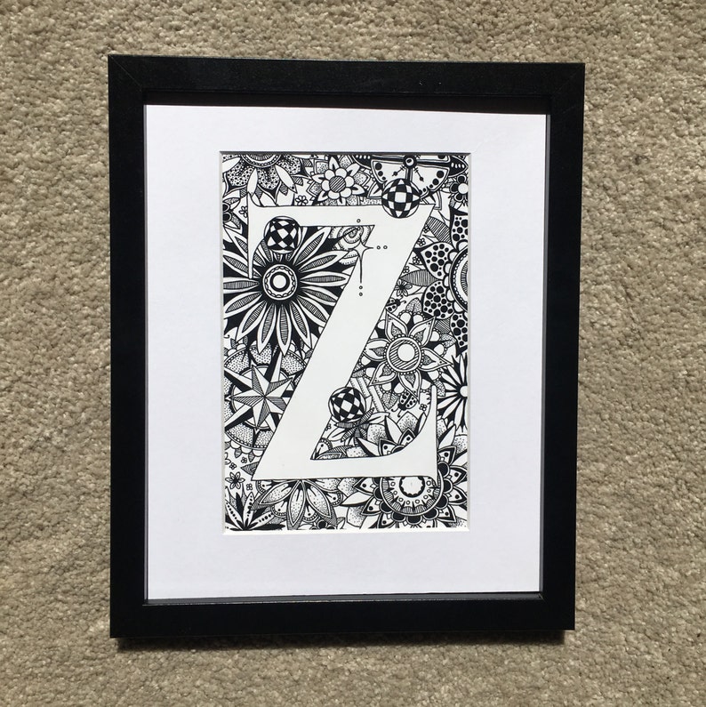 May include: A black and white framed print of the letter Z surrounded by intricate floral designs.