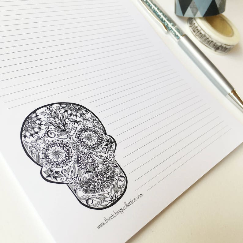 Puede incluir: Un bloc de notas blanco con l&iacute;neas y un dise&ntilde;o detallado de calavera en blanco y negro. Tambi&eacute;n se ve un bol&iacute;grafo plateado con la parte superior brillante y cinta decorativa. La direcci&oacute;n del sitio web www.theetchingscollection.com est&aacute; impresa en la parte inferior del bloc.