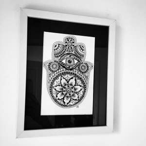 May include: A black and white framed print of a hamsa hand with an eye in the center and a mandala design. The hamsa hand is a symbol of protection and good luck.