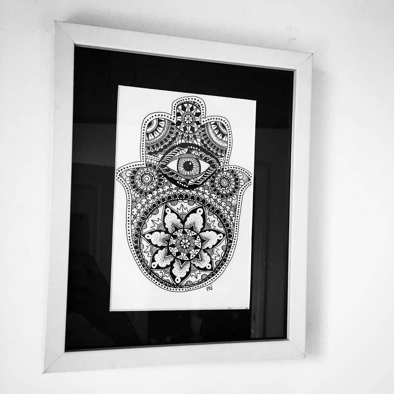 May include: A black and white framed print of a hamsa hand with an eye in the center and a mandala design. The hamsa hand is a symbol of protection and good luck.