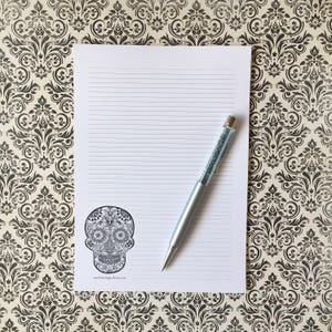 Puede incluir: Un bloc de notas blanco con l&iacute;neas y un dise&ntilde;o de calavera de az&uacute;car en blanco y negro en la esquina. Un bol&iacute;grafo plateado con un cuerpo similar a un cristal descansa sobre el bloc de notas. El fondo presenta un patr&oacute;n floral en blanco y negro.