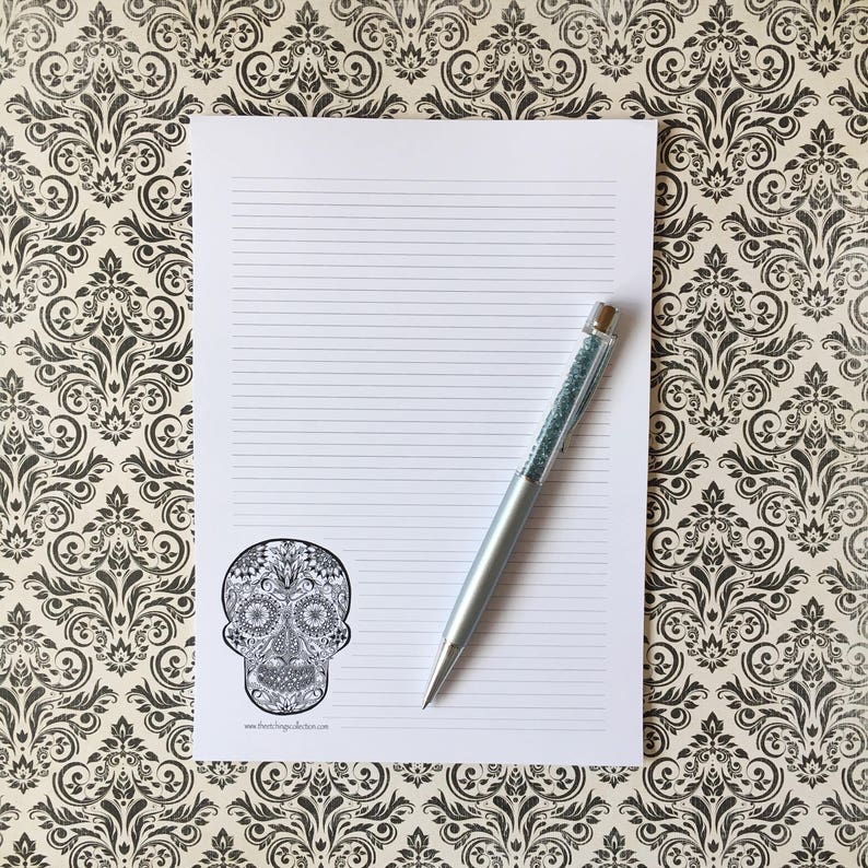 Puede incluir: Un bloc de notas blanco con l&iacute;neas y un dise&ntilde;o de calavera de az&uacute;car en blanco y negro en la esquina. Un bol&iacute;grafo plateado con un cuerpo similar a un cristal descansa sobre el bloc de notas. El fondo presenta un patr&oacute;n floral en blanco y negro.