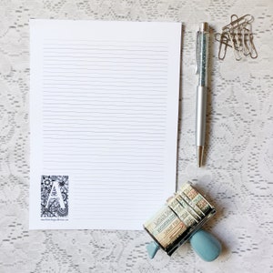 May include: A white sheet of lined paper with a black and white illustration of the letter A surrounded by flowers in the bottom left corner. A silver pen with glitter inside is on the right side of the paper. A blue and silver date stamp is in the bottom right corner.