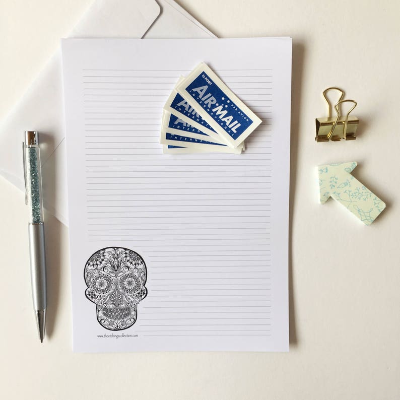 Puede incluir: Un juego de papeler&iacute;a con un dise&ntilde;o de calavera de az&uacute;car, pegatinas de correo a&eacute;reo, un bol&iacute;grafo plateado, un clip dorado y un clip en forma de flecha floral. El papel rayado est&aacute; sobre un fondo blanco.