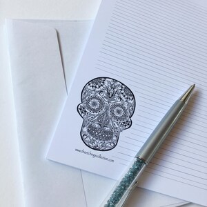 Puede incluir: Un papel de carta blanco con l&iacute;neas y una ilustraci&oacute;n detallada de una calavera en blanco y negro. Un bol&iacute;grafo plateado con detalles de cristal azul descansa sobre el papel. Un sobre blanco est&aacute; en el fondo. El sitio web www.theetchingscollection.com est&aacute; impreso.