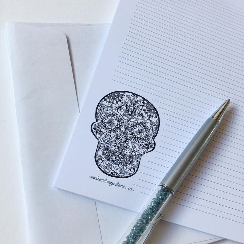 Puede incluir: Un papel de carta blanco con l&iacute;neas y una ilustraci&oacute;n detallada de una calavera en blanco y negro. Un bol&iacute;grafo plateado con detalles de cristal azul descansa sobre el papel. Un sobre blanco est&aacute; en el fondo. El sitio web www.theetchingscollection.com est&aacute; impreso.