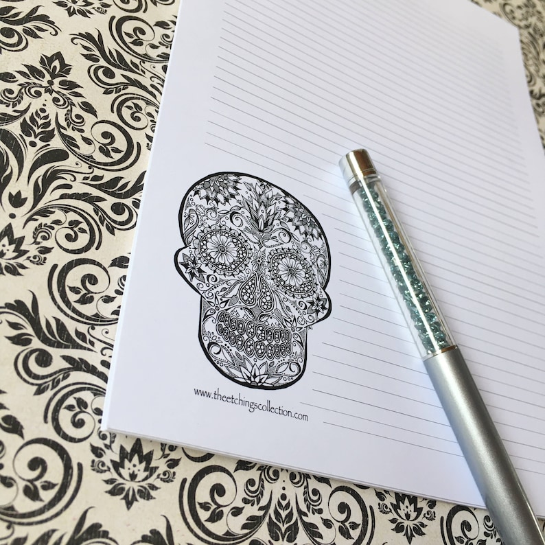 Puede incluir: Un bloc de notas rayado con un dise&ntilde;o de calavera de az&uacute;car en blanco y negro. Un bol&iacute;grafo plateado con un cuerpo lleno de cristales descansa sobre el bloc de notas. El sitio web www.theetchingscollection.com est&aacute; impreso en la parte inferior.