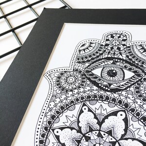 May include: A black and white hand-drawn illustration of a hamsa hand with an eye in the center. The hamsa is surrounded by intricate patterns and designs.