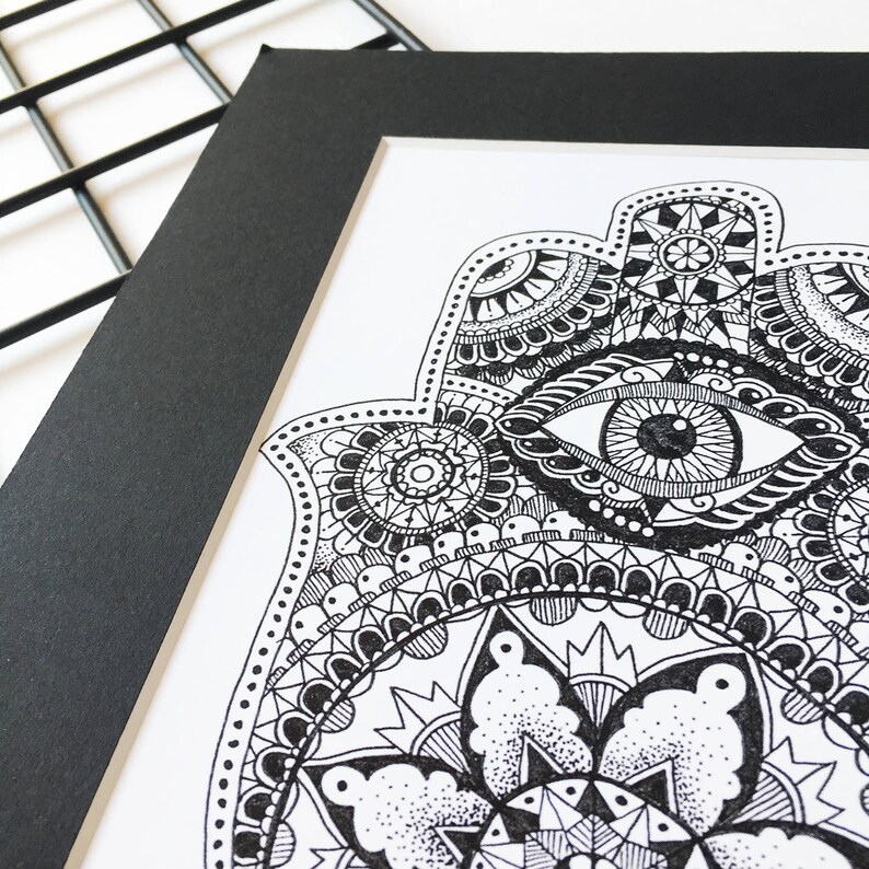 May include: A black and white hand-drawn illustration of a hamsa hand with an eye in the center. The hamsa is surrounded by intricate patterns and designs.