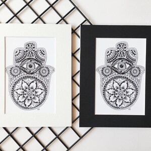 May include: Two framed prints of a black and white hamsa hand with an eye in the center and a mandala design. The frames are white and black.