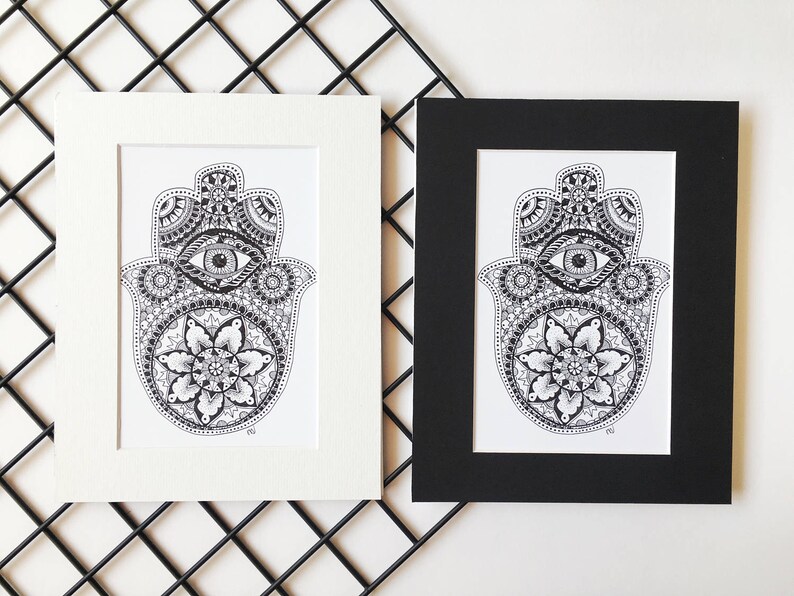 May include: Two framed prints of a black and white hamsa hand with an eye in the center and a mandala design. The frames are white and black.