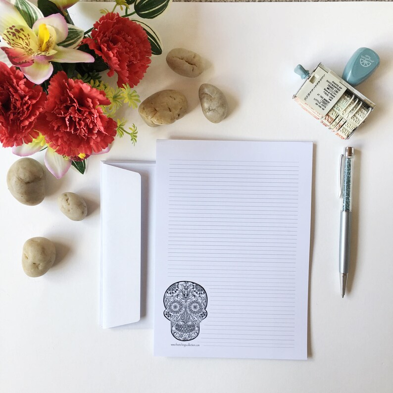 Puede incluir: Un juego de papeler&iacute;a con un bloc de notas rayado con un dise&ntilde;o de calavera de az&uacute;car en blanco y negro, un sobre y un bol&iacute;grafo con un cuerpo con incrustaciones de cristal. La imagen tambi&eacute;n incluye un sello, piedras decorativas y flores rojas.