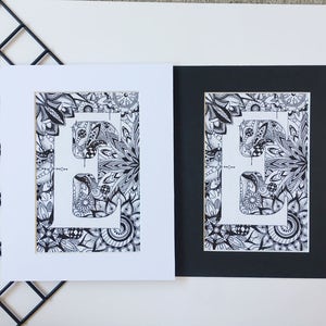 May include: Two framed prints of a black and white illustration of the letter "E" surrounded by intricate patterns. The print on the left is framed with a white mat, while the print on the right is framed with a black mat.