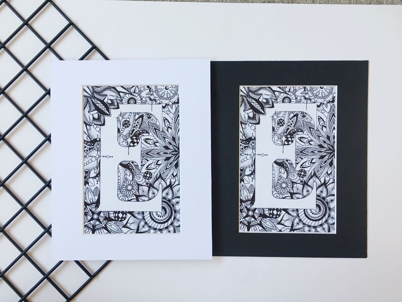 May include: Two framed prints of a black and white illustration of the letter "E" surrounded by intricate patterns. The print on the left is framed with a white mat, while the print on the right is framed with a black mat.