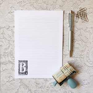 May include: A white sheet of lined paper with a black and white illustration of the letter "B" in the lower left corner. A silver pen with glitter inside is on the right side of the paper. A blue and silver stamp with various numbers and letters is in the lower right corner.