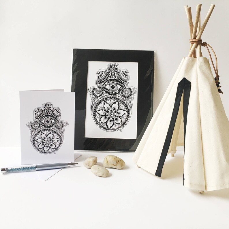 May include: A black and white illustration of the Hamsa hand, a symbol of protection, with intricate details and a central eye. The illustration is printed on a white card and framed in a black frame. The card is displayed next to a small teepee made of white fabric with black trim.