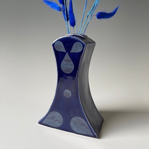 May include: A small, dark blue ceramic vase with a geometric design. The vase has a wide base and a narrow neck. It is decorated with white geometric shapes, including circles and teardrops. The vase is filled with blue dried flowers.