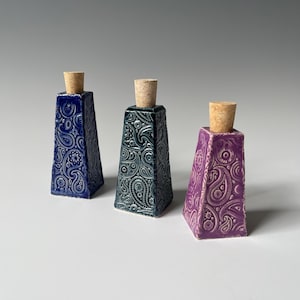 Könnte beinhalten: Drei Keramikflaschen mit konischer quadratischer Form und glänzender Oberfläche. Jede Flasche hat ein erhabenes Paisley-Muster und ist mit einem Naturkorken verschlossen. Die Flaschen sind in den Farben Dunkelblau, Dunkeltürkis und Violett gehalten.