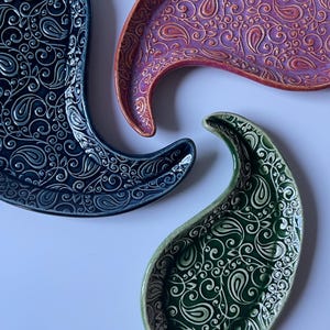 Puede incluir: Primer plano de tres fuentes de cerámica con forma de paisley. Las fuentes son de color azul marino, morado y verde, cada una con un diseño en relieve de paisley y volutas. Las fuentes están sobre un fondo blanco.
