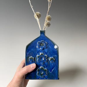 May include: A small, blue ceramic vase with a unique, arched shape and embossed flower designs. The vase is perfect for displaying a single stem or small bouquet of flowers.