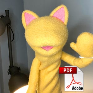 May include: A yellow plush hand puppet with pointed ears featuring pink inner accents and a pink mouth. The puppet has a rounded head and a raised arm. A white plastic tag is attached to the arm of the puppet, which is positioned near a lamp.