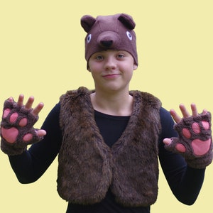 Op de afbeelding: Een kind dat een bruin berenpak draagt met een bontvest, een bruine hoed met berenoren en roze pootjeshandschoenen.