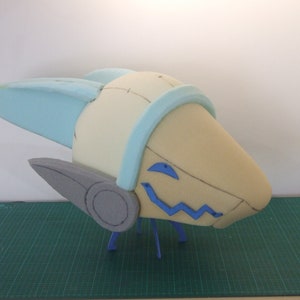 May include: A foam sculpture of a head with blue and gray accents. The head has a large, rounded shape with two small, pointed ears on the sides. The head is decorated with blue and gray accents, including a blue band around the top of the head and a gray circle on the side of the head. The head is sitting on a green surface.