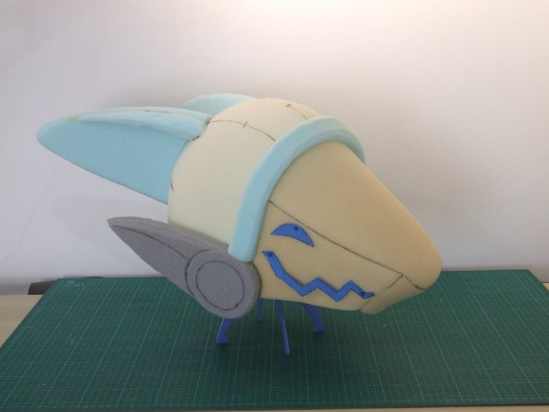 May include: A foam sculpture of a head with blue and gray accents. The head has a large, rounded shape with two small, pointed ears on the sides. The head is decorated with blue and gray accents, including a blue band around the top of the head and a gray circle on the side of the head. The head is sitting on a green surface.