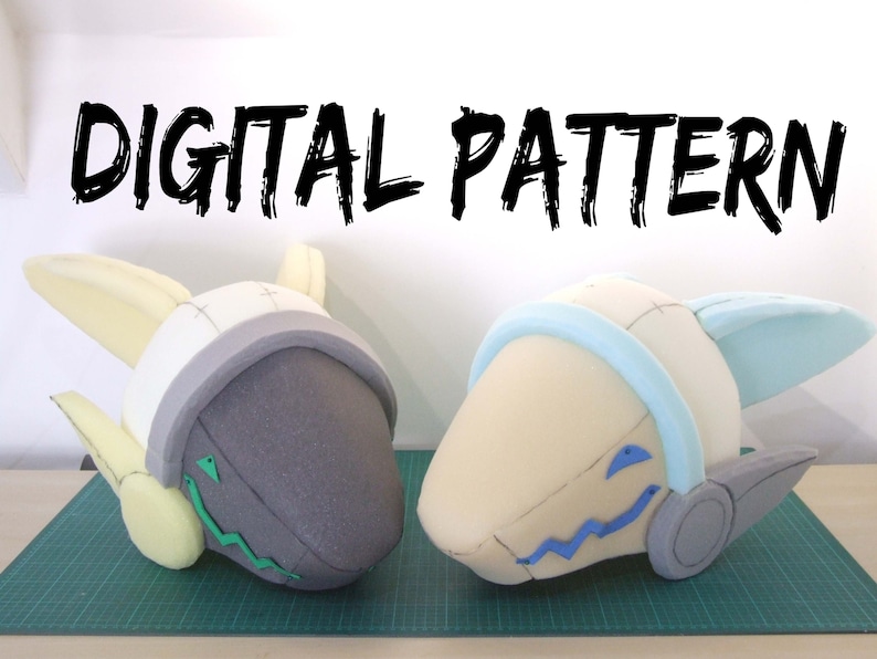 May include: Two foam helmet forms with details drawn on them. The helmet on the left is gray with green details and yellow ears. The helmet on the right is white with blue details and light blue ears. The helmets are on a green cutting mat. The text "DIGITAL PATTERN" is written in black at the top of the image.