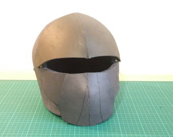 May include: A dark gray helmet with a rounded top and a face opening. The helmet appears to be made of a rigid material and has visible seams. It is placed on a green grid surface.