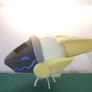 May include: A white foam model of a spaceship with yellow wings and a gray and black head. The model is on a green cutting mat and has white legs.