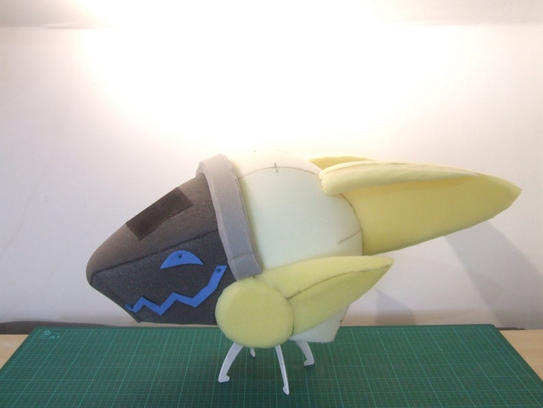 May include: A white foam model of a spaceship with yellow wings and a gray and black head. The model is on a green cutting mat and has white legs.