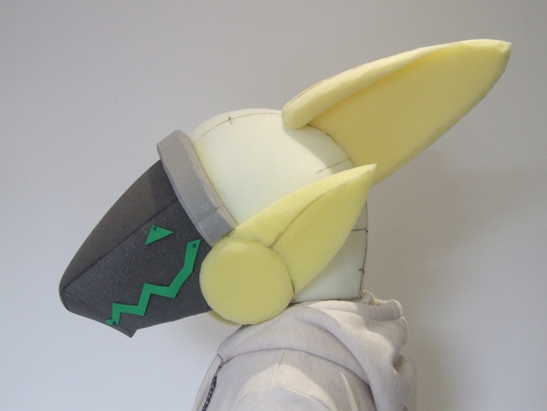 May include: A foam head with a gray and green face mask and yellow foam ears. The head is on a white mannequin.