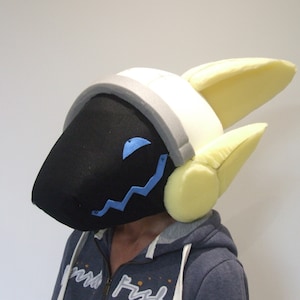 May include: A black and white furry head with yellow ears and a gray headband. The head has a blue zig-zag design on the front and a blue eye.