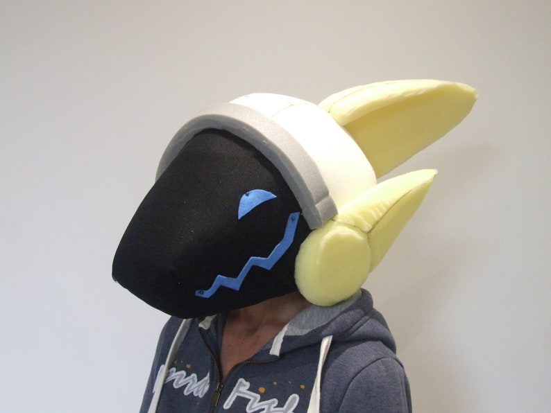 May include: A black and white furry head with yellow ears and a gray headband. The head has a blue zig-zag design on the front and a blue eye.