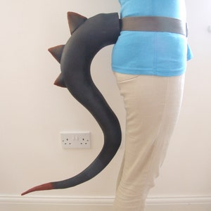May include: A black and red dragon tail costume accessory. The tail is made of foam and has a textured surface. It is attached to a belt and can be worn around the waist.