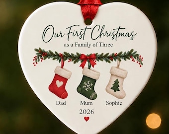 May include: A white heart-shaped ceramic ornament hanging from a red ribbon. It features the text Our First Christmas as a Family of Three, a green garland with red berries, and three stockings labelled Dad, Mum, and Sophie with the year 2026.