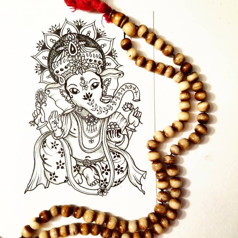 Puede incluir: Un dibujo en blanco y negro de la deidad hind&uacute; Ganesha, con cabeza de elefante, sentado en una postura meditativa. Un mala de madera marr&oacute;n, o cuentas de oraci&oacute;n, est&aacute; drapeado sobre el dibujo.