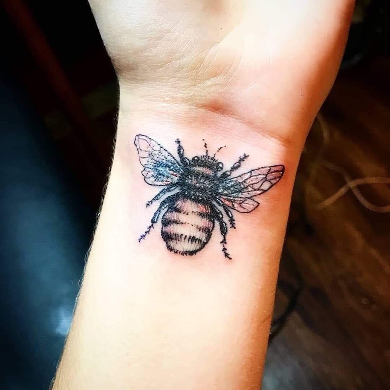 Puede incluir: Un tatuaje en blanco y negro de una abeja en la mu&ntilde;eca de una persona.