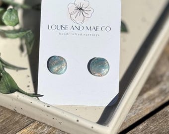 May include: A pair of handcrafted stud earrings with a light blue and gold glitter design. The earrings are displayed on a white card with the text "LOUISE AND MAE CO handcrafted earrings". The earrings are round and sit on a white tray.